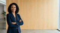 Young woman with curly dark hair and medium skin tone wearing navy blazer, arms crossed, smiling confidently in modern office with wooden wall background.