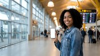Young African American woman holding blank smartphone screen at modern airport terminal with departure boards and travelers in background.
