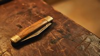 Vintage wooden folding pocket knife with brass fittings resting on aged leather surface with embossed map pattern creating rustic antique atmosphere.