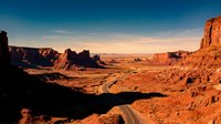 Scenic desert highway stretches through vast arid landscape with towering red rock formations and buttes under golden sunset sky, suitable for travel and nature backgrounds.