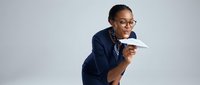 Professional African American businesswoman in navy suit throwing paper airplane on gray background for corporate creativity concepts.