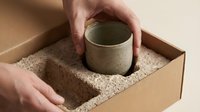 Hands carefully unpacking handmade ceramic mug from protective cardboard box with eco-friendly packaging materials for safe shipping delivery.