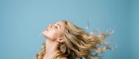 Young blonde woman with flowing hair in motion against blue background expressing joy and freedom with head tilted back and eyes closed in carefree moment.