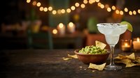 Margarita cocktail with lime garnish served alongside guacamole and tortilla chips on wooden table with warm string lights and candles in background for entertaining and celebration occasions.