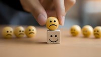 Hand placing sad yellow emoji face on wooden cube with happy face drawing, surrounded by unhappy emoji balls on wooden table. Concept of mood change.