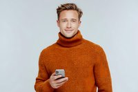 Young Caucasian man in orange turtleneck sweater holding smartphone against light gray background, smiling confidently at camera.