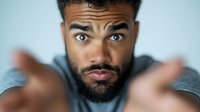 Young African American man with beard and intense gaze reaching toward camera with blurred hands in foreground, expressing surprise and curiosity against light blue background.