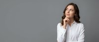 Young professional woman in white shirt looking up thoughtfully with hand on chin, contemplating future possibilities on gray background.