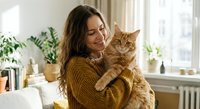 Young woman cuddling fluffy orange tabby cat in cozy home interior with plants. Pet owner bonding moment for lifestyle and domestic comfort concepts.