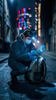 Young Asian man wearing face mask crouching on urban street at night using smartphone with colorful city lights bokeh background creating atmospheric mood.
