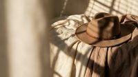 Brown leather cowboy hat with decorative band resting on white fabric in warm sunlight creating dramatic shadows for western fashion and lifestyle concepts.
