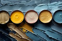 Colorful oil paint jars with thick textured paint in yellow, pink, brown and blue tones arranged in row on dark artistic background with palette knife strokes.