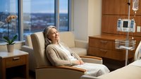 Pregnant woman relaxing in comfortable hospital chair during medical treatment with IV drip in modern healthcare facility room with city view.