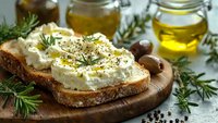 Fresh goat cheese spread on rustic bread with herbs and olive oil, Mediterranean appetizer setup on wooden board with rosemary garnish and olives.