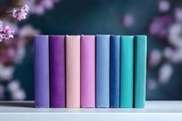 Row of colorful hardcover books arranged in rainbow gradient from purple to turquoise on white surface with soft pink cherry blossoms bokeh background.