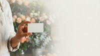 Young adult male with medium skin tone wearing white casual shirt holding blank business card outdoors with blurred garden background for branding and marketing purposes.