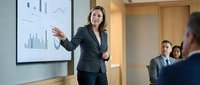 Professional businesswoman presenting financial data charts to corporate team in modern conference room during strategic meeting.