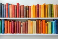 Colorful vintage hardcover books arranged on white wooden shelves, spines showing rainbow spectrum from blue through red to yellow and green. Library collection display.