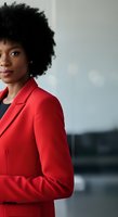 Young African American woman with natural curly hair wearing a bright red blazer over dark top, standing confidently against blurred urban background for corporate and business use.