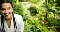 Young woman with braided hair and olive complexion wearing white blazer smiling outdoors in lush garden setting for business, lifestyle, and wellness content.
