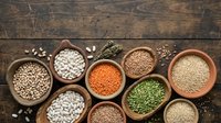 Assorted legumes and grains in clay bowls on rustic wooden surface, including lentils, chickpeas, beans and seeds for cooking and nutrition.