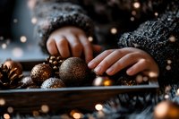 Female hands arranging copper and bronze Christmas ornaments on dark surface with bokeh lights and pine cones creating cozy festive atmosphere.