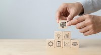 Businessman hands arranging wooden blocks with target and business icons on desk for strategic planning and goal achievement in corporate environment.