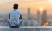 Young Asian man in casual wear sitting alone on rooftop edge overlooking city skyline during golden sunset, back view capturing contemplative solitary moment.