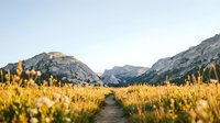 Young woman with long blonde hair, wearing casual outdoor clothing, standing in a golden meadow with mountain landscape in background during golden hour, suitable for travel, lifestyle, and nature editorial use.