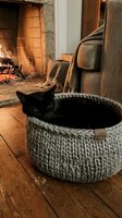Black cat resting in knitted basket near fireplace with cozy blankets and warm ambiance, perfect for home decor and pet lifestyle content.