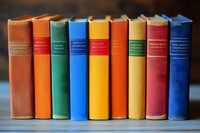 Row of colorful vintage hardcover books standing on wooden shelf, spines showing with blurred background, arranged in rainbow spectrum order.