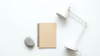 Minimalist workspace with kraft paper notebook, white desk lamp and smooth stone on clean white background for modern office design and productivity concepts.