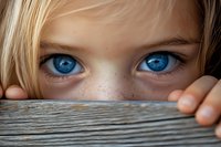 Close up view of bright blue eyes and freckled nose peeking over wooden surface, showing curiosity and playful hiding expression. Blonde hair visible.