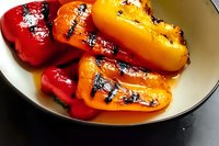 Grilled red and yellow bell peppers with charred grill marks and glossy surface on white ceramic plate, close up view showing rich colors and roasted texture.