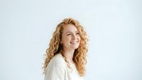 Middle-aged woman with curly blonde hair wearing white casual shirt, smiling while looking away against neutral background. Portrait suitable for business, lifestyle, and wellness content.