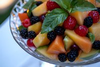 Fresh summer fruit salad with cantaloupe chunks, blackberries, raspberries and basil leaves in crystal bowl, macro food photography on light background.