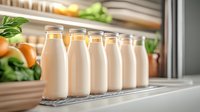 Fresh dairy bottles with creamy milk lined up in refrigerator door shelf, organic produce and vegetables visible in background, healthy food storage concept.
