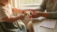 Child giving small flower bouquet to adult woman for Mother's Day celebration at home with handmade card on wooden table in warm natural light.
