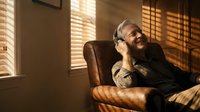 Relaxed middle-aged Caucasian man with beard enjoying music through headphones while sitting in leather armchair with warm sunlight streaming through blinds.