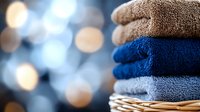 Stack of neatly folded fresh bath towels in blue and gray colors on wicker basket against blurred bokeh lights background, clean laundry concept.