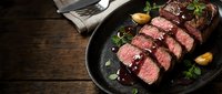 Sliced medium-rare beef steak with herbs and sauce in cast iron skillet on rustic wooden table. Gourmet restaurant meal ready to serve.