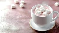 White cup of pink hot chocolate with marshmallows on saucer against soft pink background with scattered marshmallows and powdered sugar dusting.