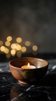 Ceramic bowl with burning candle on marble surface creating warm ambiance with golden bokeh lights in background for spa and relaxation concepts.