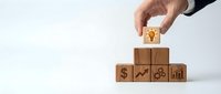 Hand placing wooden cube with lightbulb icon on top of business blocks with dollar, chart, gear and graph symbols representing innovation and growth strategy.