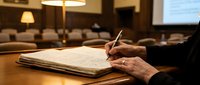 Professional businessman writing notes in leather notebook during corporate meeting in modern conference room with warm lighting and blurred colleagues.
