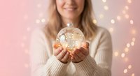 Young blonde woman holding glowing snow globe with warm lights on pink background. Holiday magic and winter celebration concept for greeting cards.