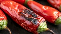 Grilled red chili peppers with charred skin and vibrant green stems on dark background, macro food photography showing texture and grill marks.