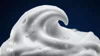 White creamy foam wave splash with smooth texture and droplets against dark blue background for cosmetic and skincare product advertising.
