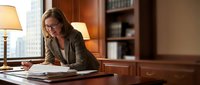 Professional businesswoman with glasses working at wooden desk in elegant office interior with warm lighting and bookshelves reviewing documents.