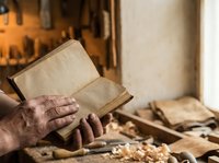 Craftsman holding open aged book surrounded by wood shavings and carving tools in workshop. Suitable for woodworking, craftsmanship, and artisan content.